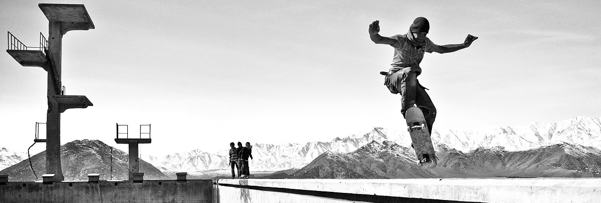The picture shows skateboarders who perform daring stunts in a disused swimming pool and face a mountain panorama.